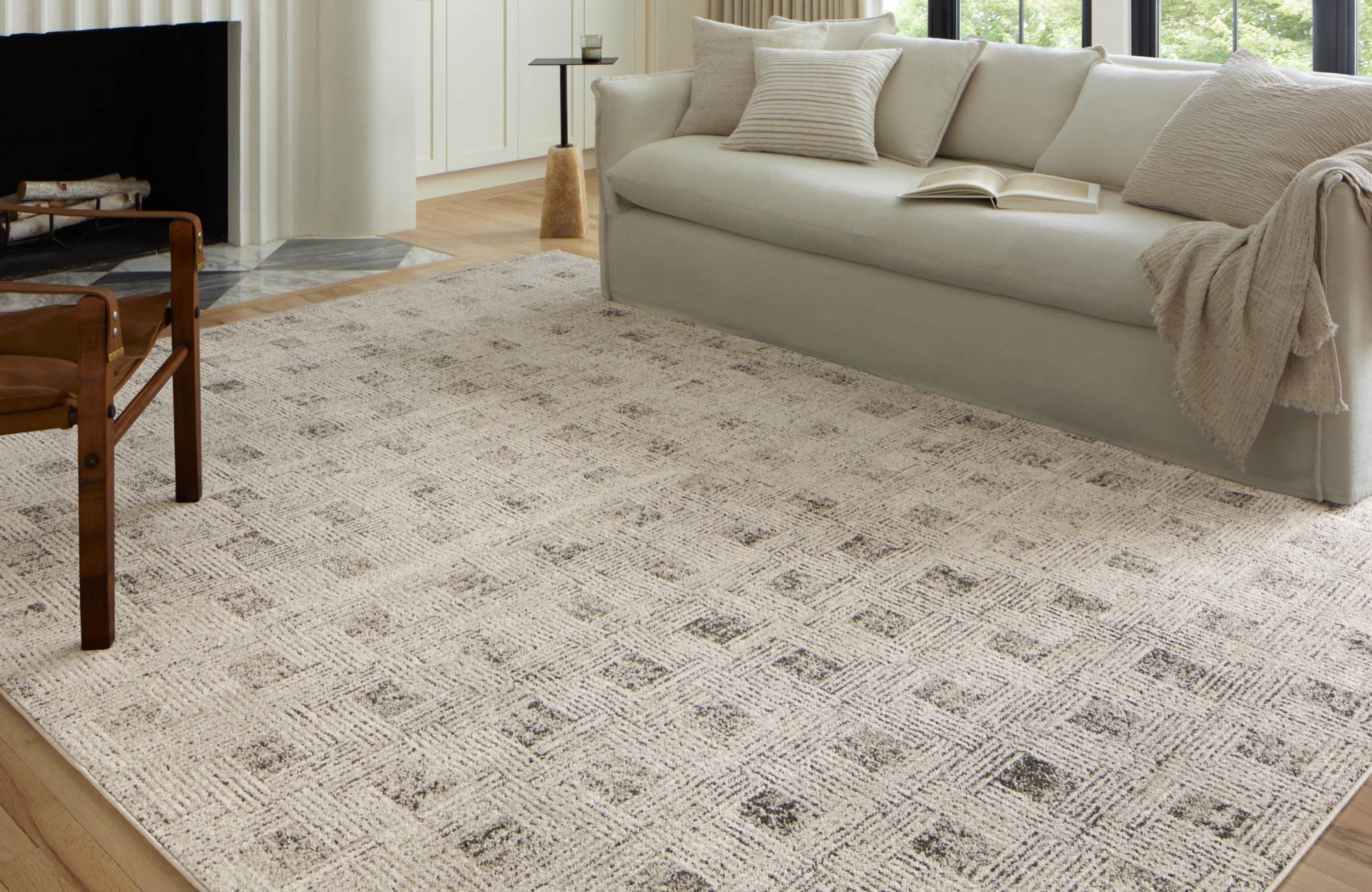 Neutral patterned area rug grounding a soft-toned living room with a long sofa, modern chair, and open fireplace.