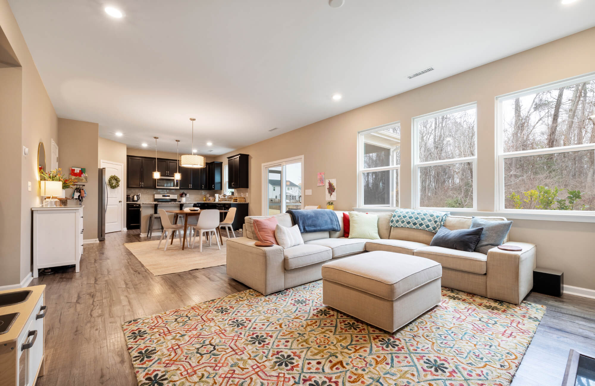 Open-concept living room and kitchen with beige sectional, colorful patterned rug, neutral walls, and large windows bringing in natural light.