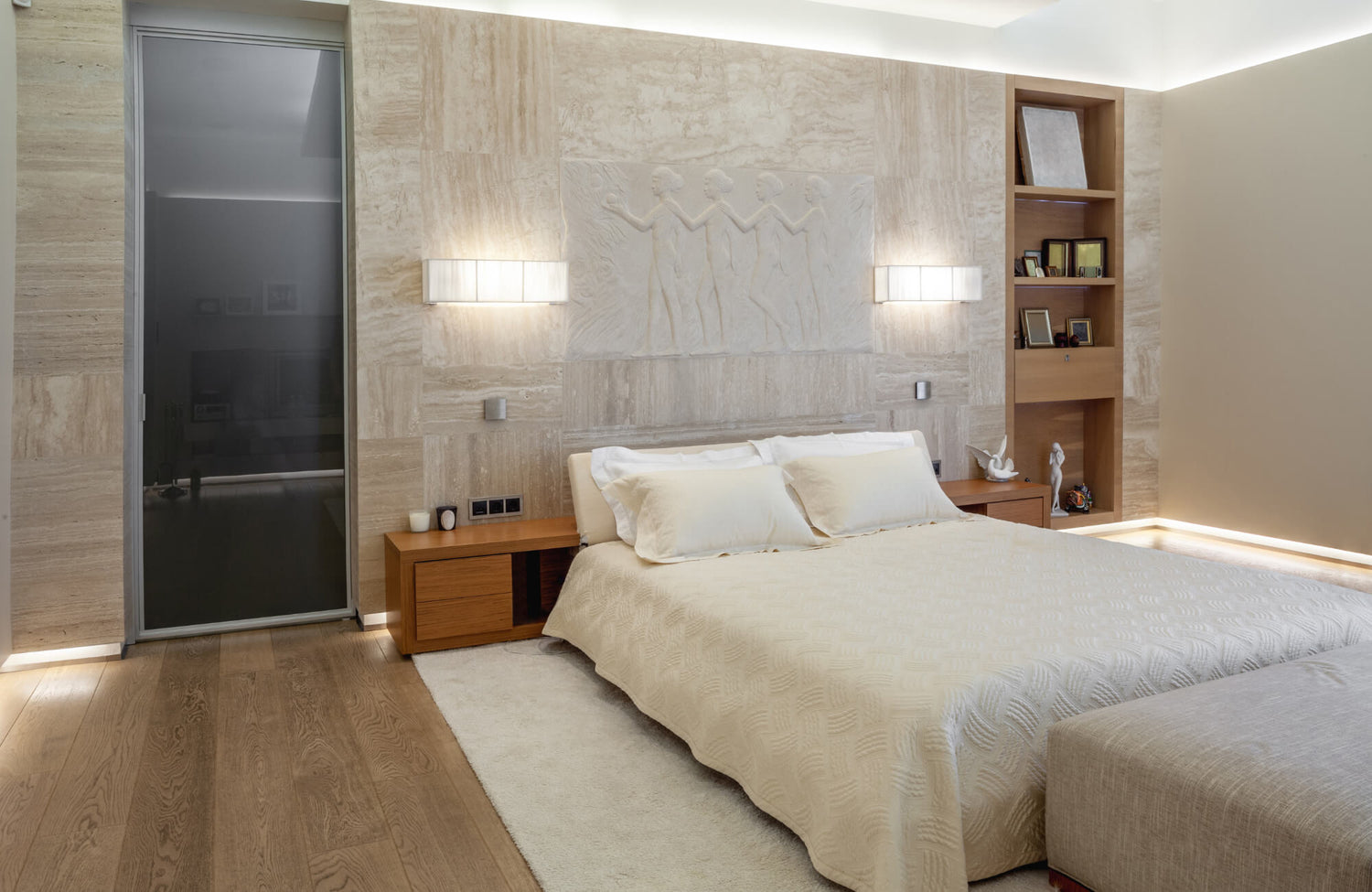 Modern bedroom with warm wood accents, recessed lighting, and symmetrical wall sconces flanking a sculpted stone headboard feature.