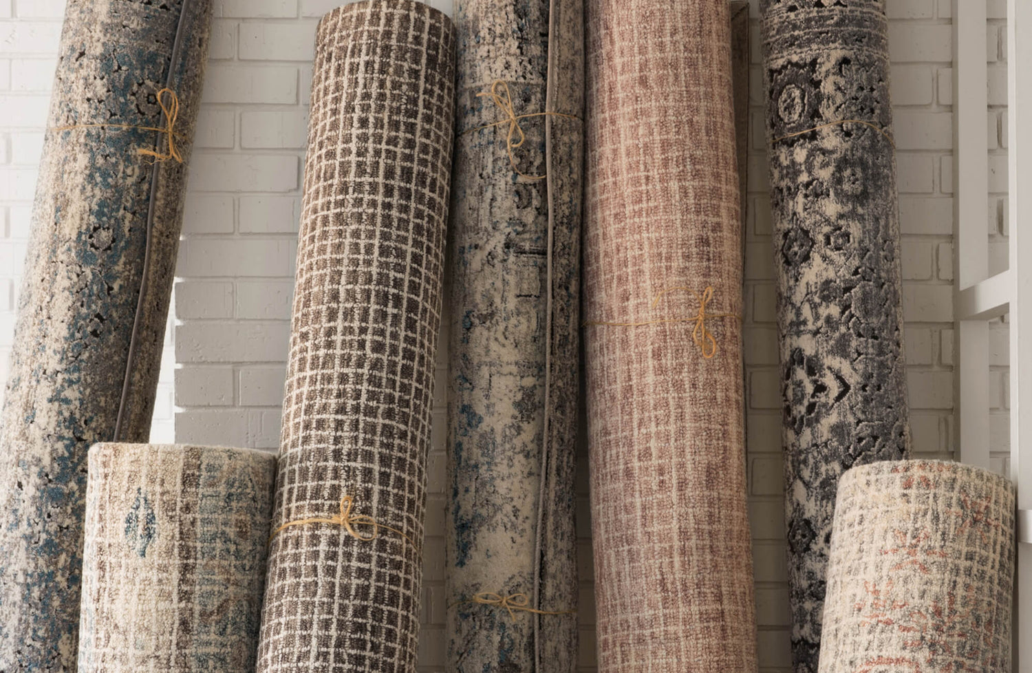Rolled high-quality area rugs in earthy tones and distressed patterns leaning against a white brick wall, showcasing vintage-inspired textures.
