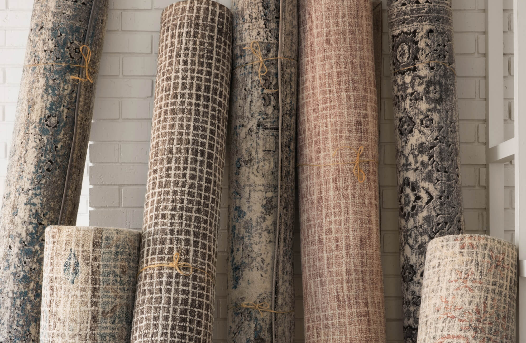 Rolled high-quality area rugs in earthy tones and distressed patterns leaning against a white brick wall, showcasing vintage-inspired textures.