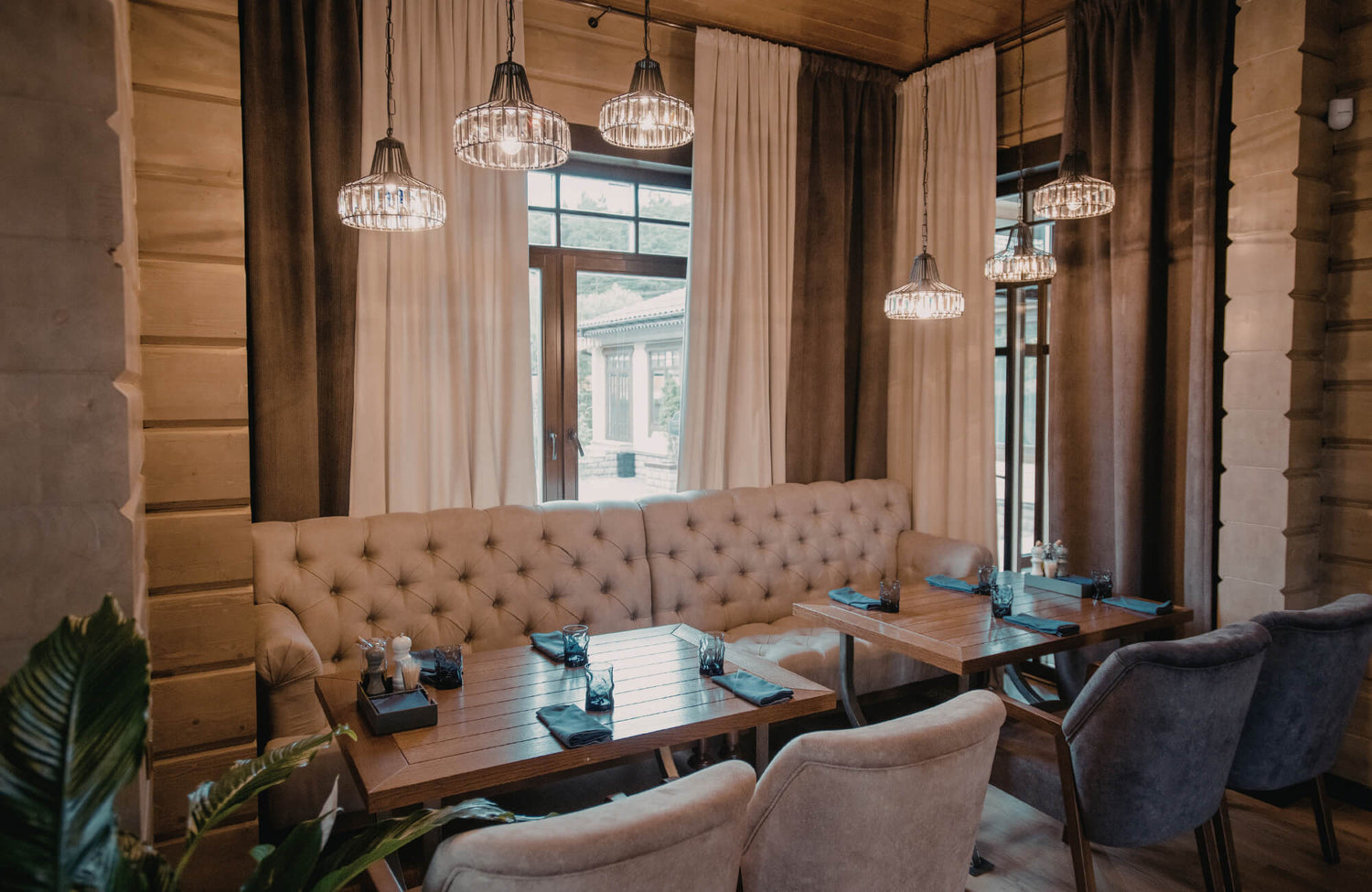 Glass pendant lights cast warm ambient light over a cozy dining banquette with wood tables, soft upholstery, and floor-to-ceiling curtains.