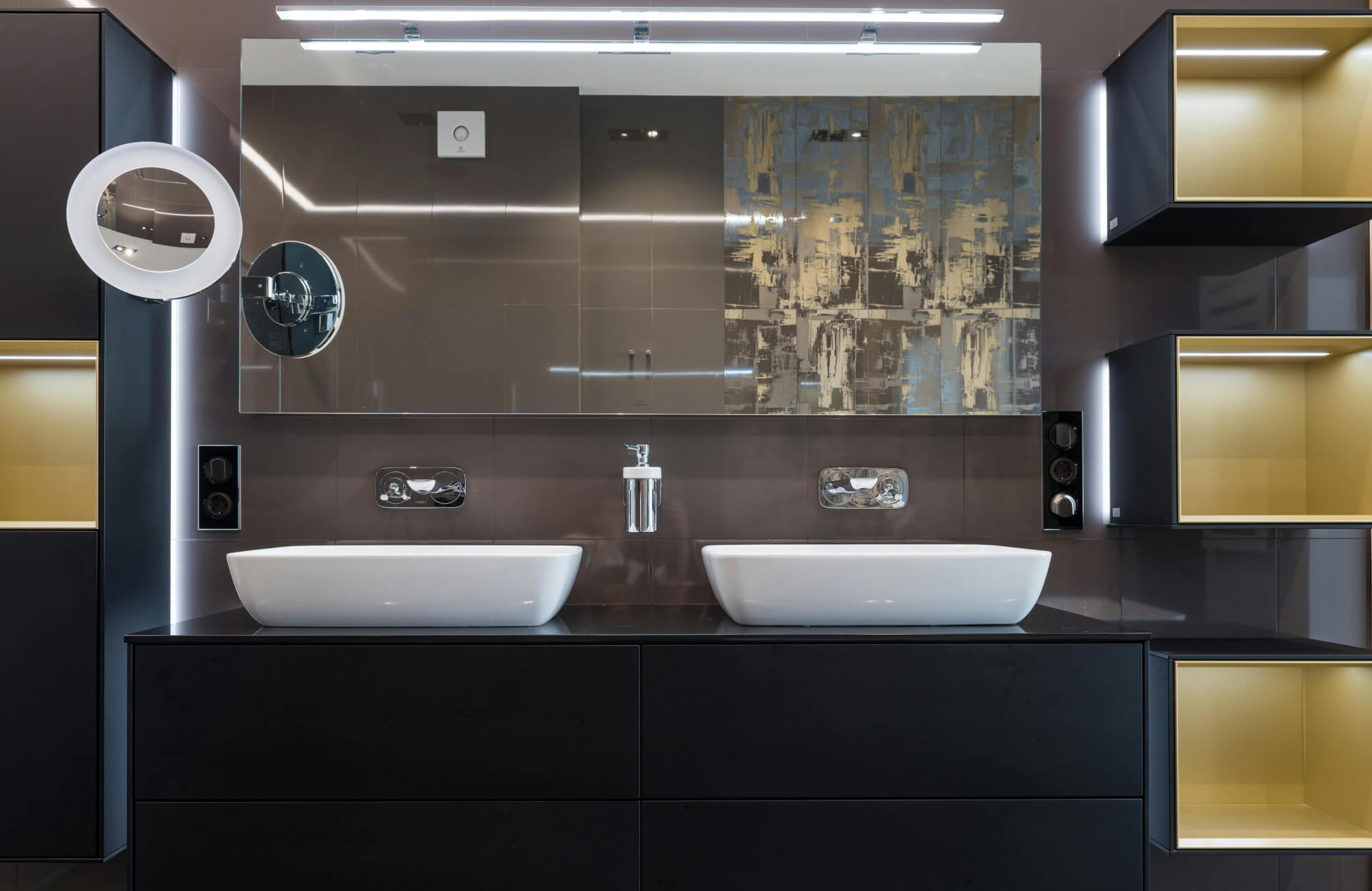 A modern dining room set featuring a round matte-black pedestal table, plush taupe upholstered chairs with gold-tipped legs, and a soft, moody backdrop creates an elegant, warmly lit space anchored by a sculptural floral centerpiece.