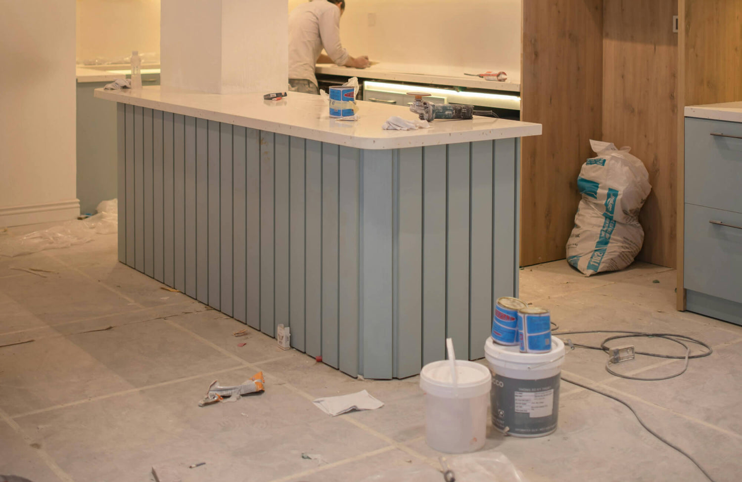 Modern kitchen under renovation featuring a light blue vertical panel island with a white quartz countertop, wood cabinetry, and tools scattered across the unfinished tiled floor.