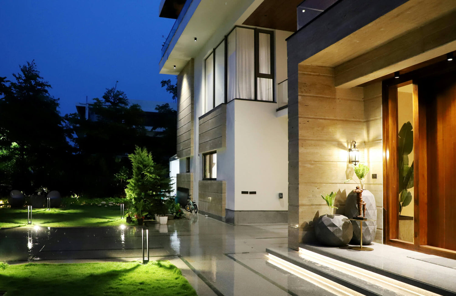 Modern home exterior at dusk with large format stone tile entry, illuminated steps, a warm wall sconce, wood accents, and landscaped greenery.