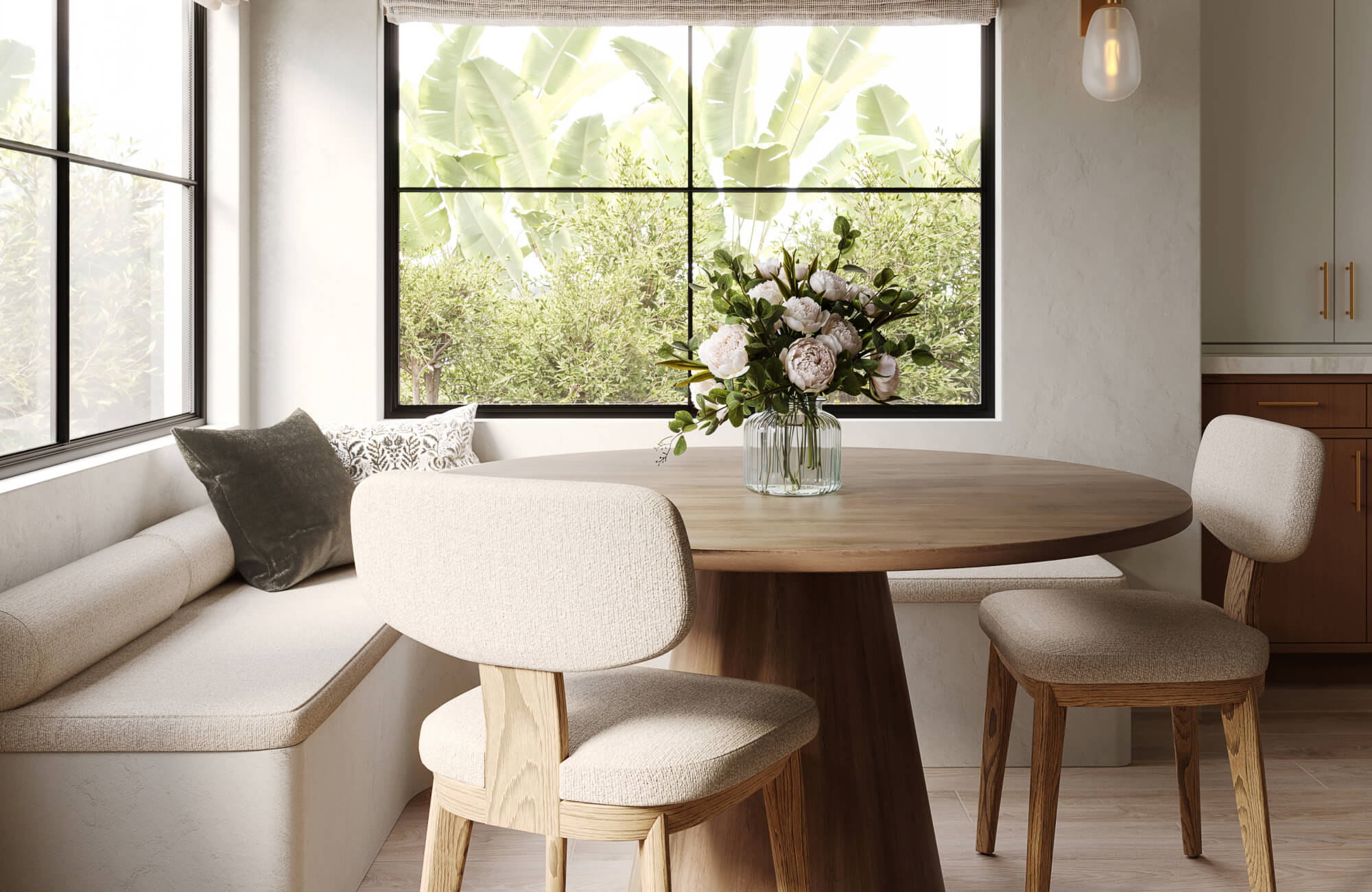 Cozy breakfast nook with a round wooden table, upholstered seating, and a glass vase of peonies under sunlit windows.