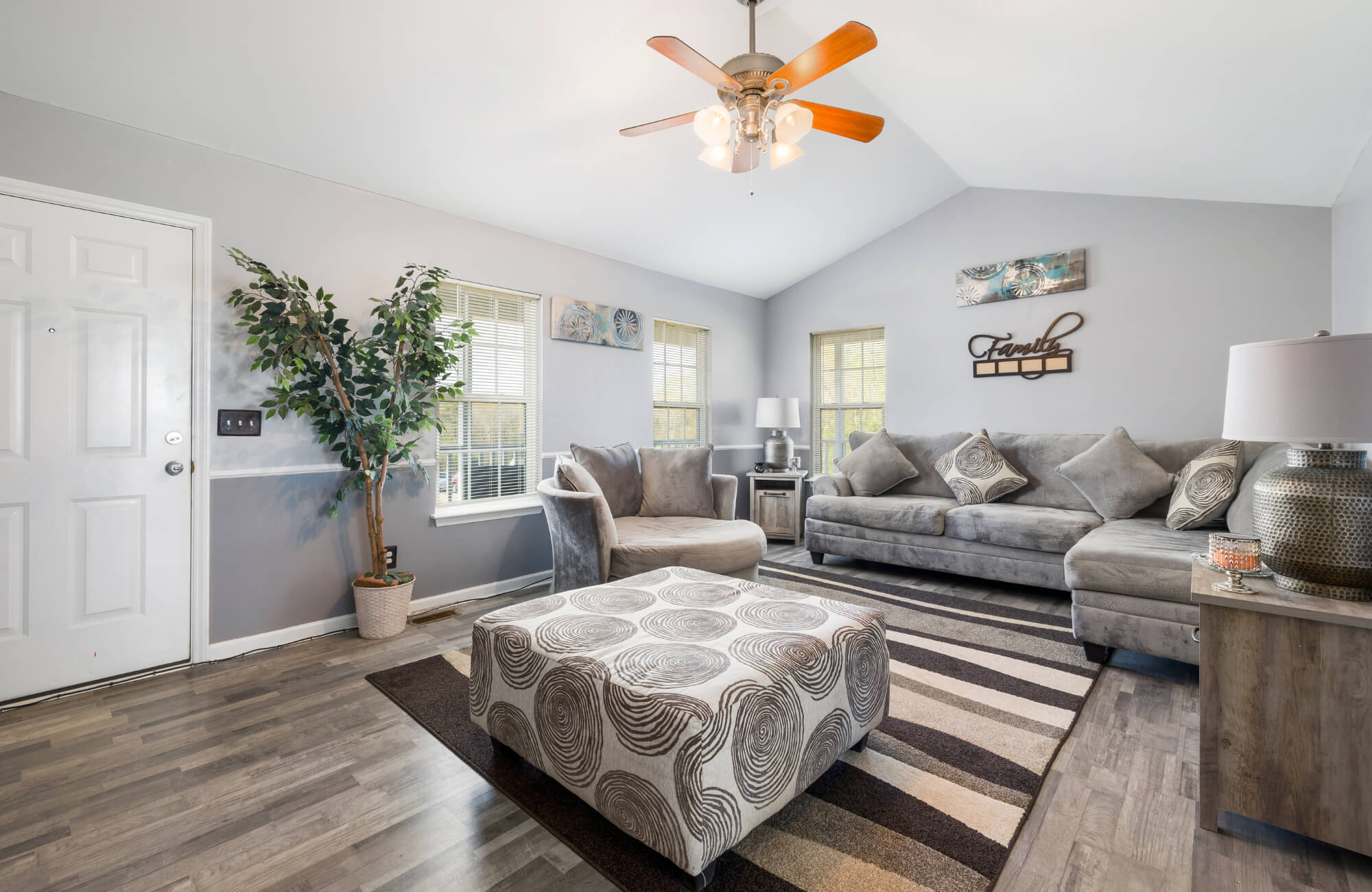 Modern living room with gray furniture, striped area rug, wood flooring, and neutral decor accents under natural and ceiling light.