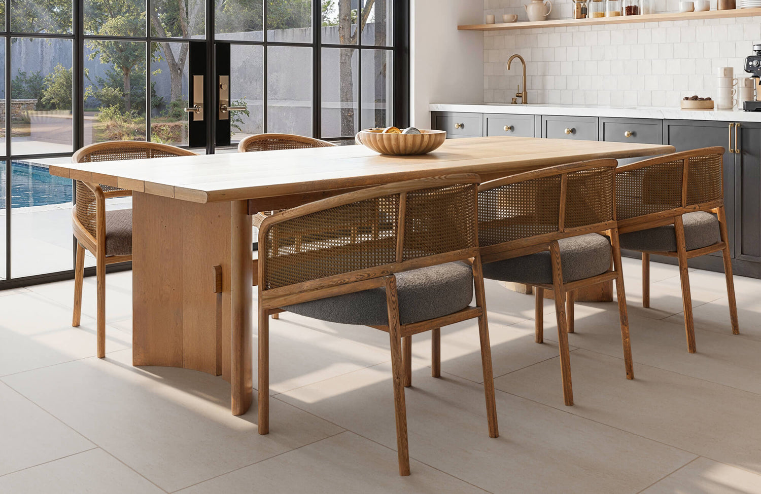 Sunlit modern kitchen showcasing a solid wood dining table with woven-back chairs, soft gray cabinetry, white tiled backsplash, and expansive black-framed glass doors.