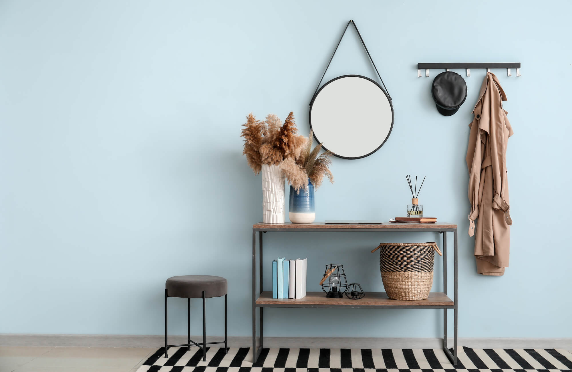 Entryway with a round mirror, wooden console table, decorative vases, coat rack, and neutral-toned decor against a light blue wall.