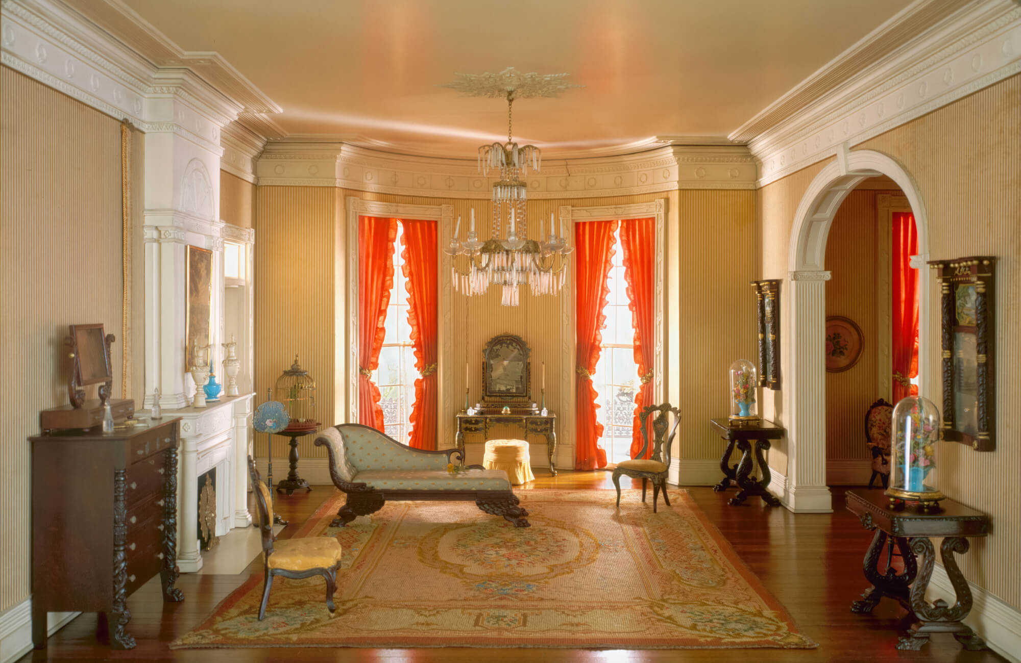 Elegant parlor with dark wood floors, ornate Victorian furniture, bold red drapes, and a large orange floral rug.