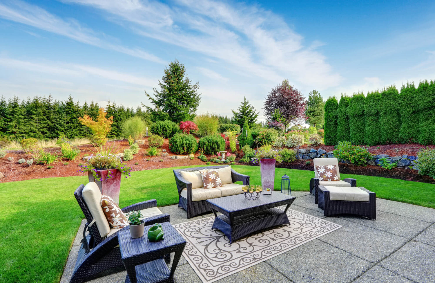 Outdoor patio with wicker furniture, neutral cushions, a decorative rug, and a manicured garden with lush greenery and colorful shrubs.