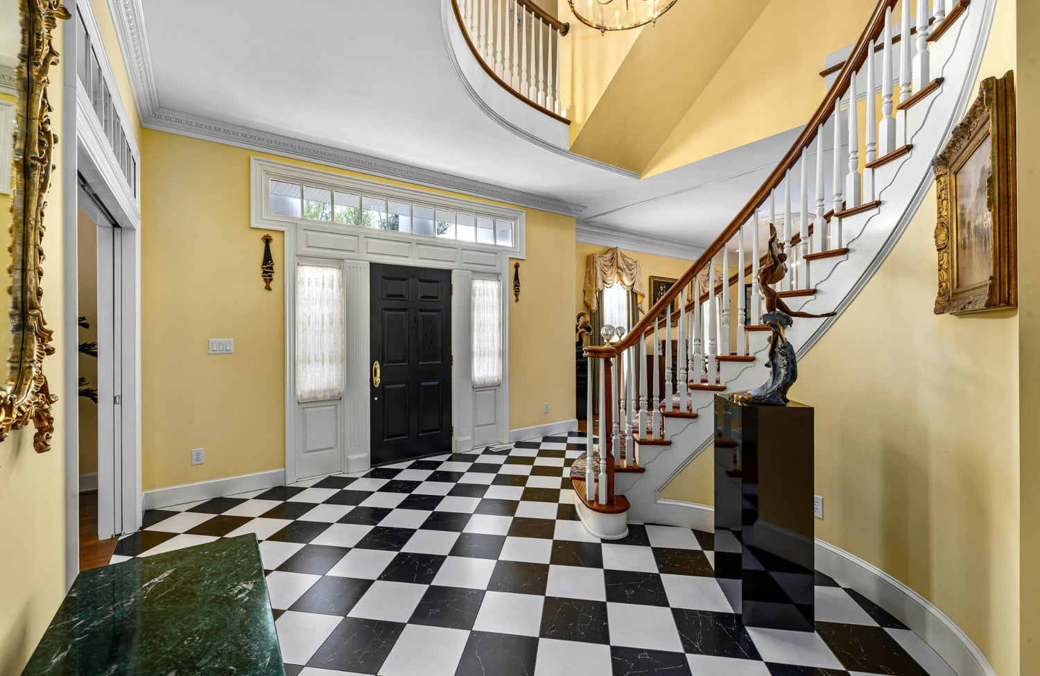 Elegant foyer with a black-and-white checkerboard marble floor, a grand curved staircase, yellow walls, and a black double-door entrance.