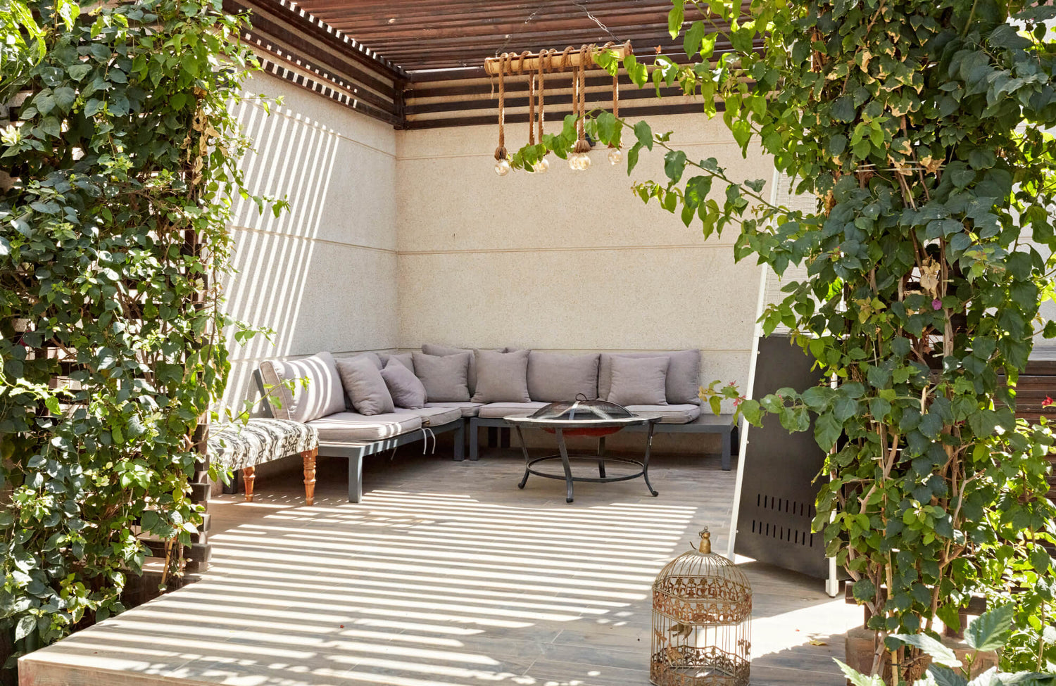 A shaded patio with cushioned seating, wood pergola slats, and lush climbing greenery creating a cozy, inviting outdoor lounge.