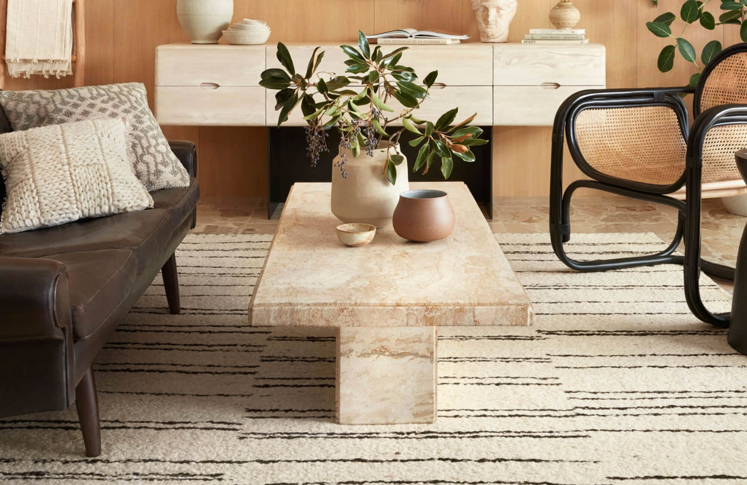 Modern living room with a striped cream and black rug, leather sofa, rattan chair, and a travertine coffee table topped with ceramics.