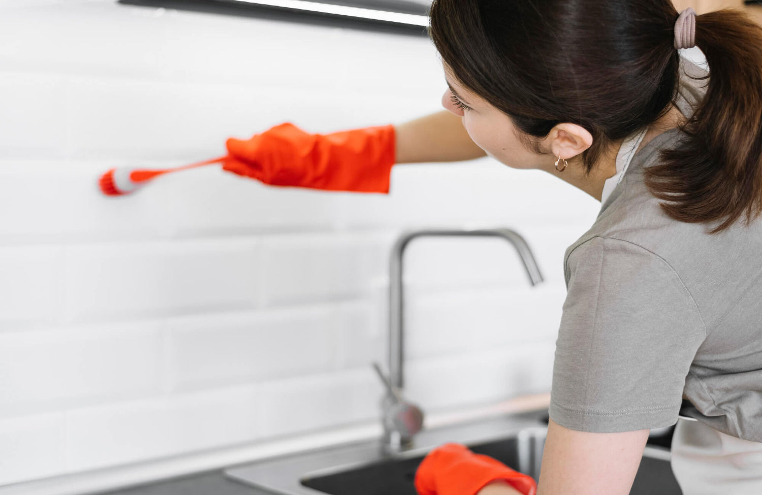 A woman wearing orange cleaning gloves scrubs the white subway tile backsplash above a kitchen sink using a small brush.