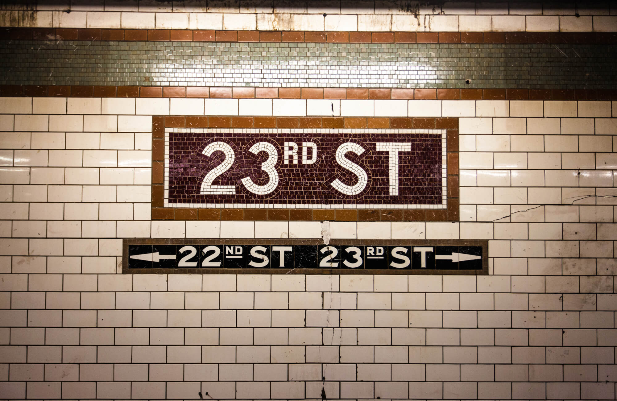 Vintage subway wall with white ceramic tiles and a mosaic sign reading “23rd ST,” showing grime, cracks, and directional street markers.