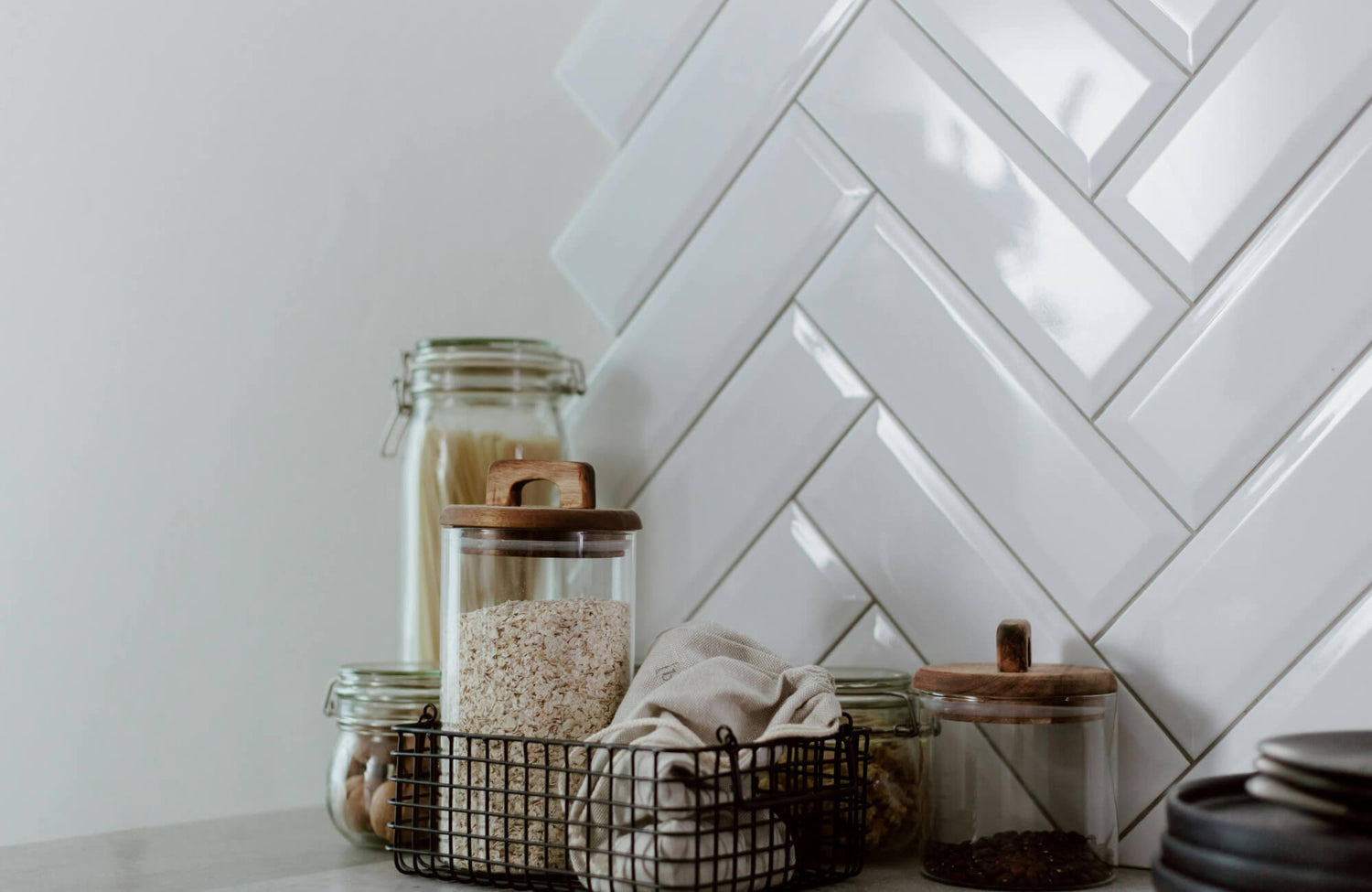 Glossy white herringbone subway tile backsplash in a pantry, paired with glass jars, wooden lids, and a black wire storage basket.