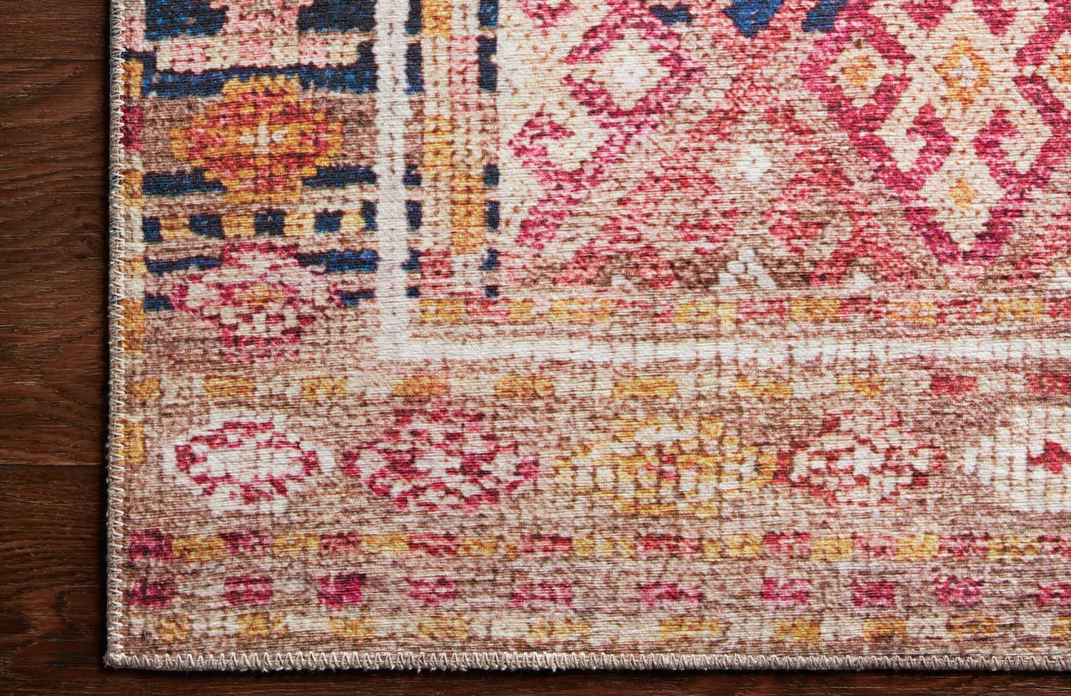 Close-up of a rug corner with faded pink, gold, and muted blue geometric motifs, finished with a stitched edge on dark wooden flooring.