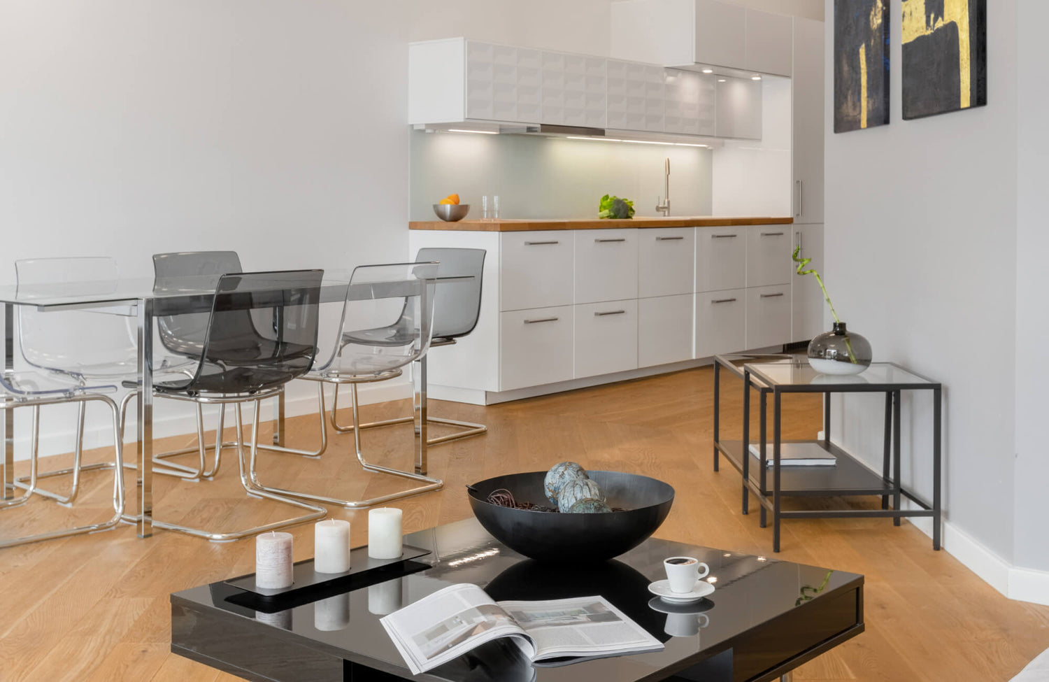 Modern dining area with a glass table and transparent chairs beside a sleek white kitchen and warm wood flooring.
