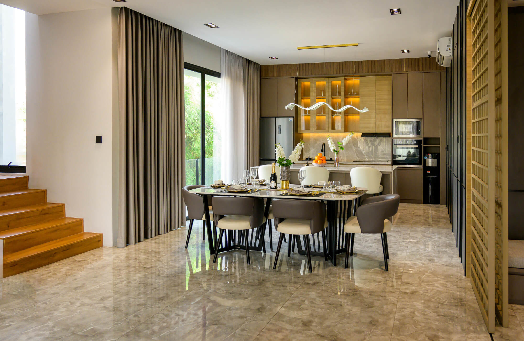 Modern open-plan dining area with a long rectangular table, mixed upholstered chairs, warm lighting, and a sleek kitchen backdrop.