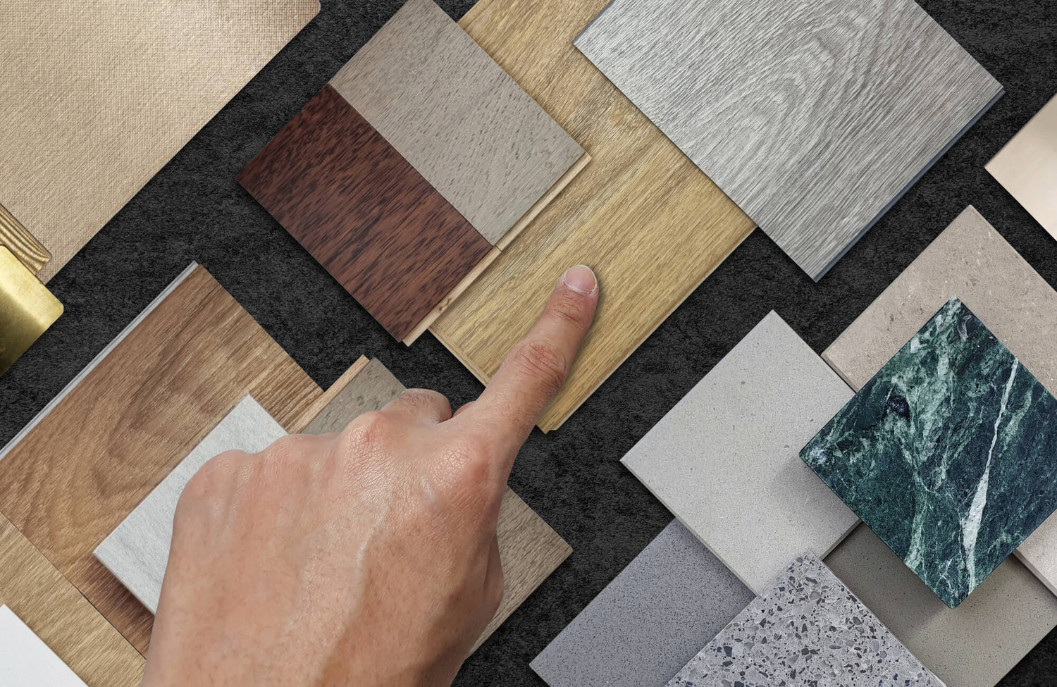 A hand selecting a wood flooring sample from an assortment of wood, stone, and tile swatches, arranged on a dark textured surface.