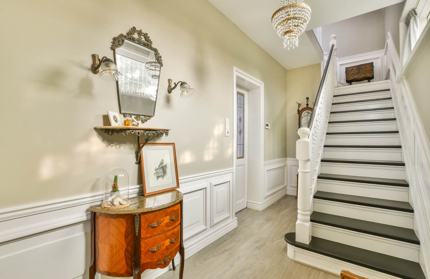 Vintage hallway with ornate wall mirror, carved sconces, wood chest, and a white staircase with wainscoting and a chandelier.