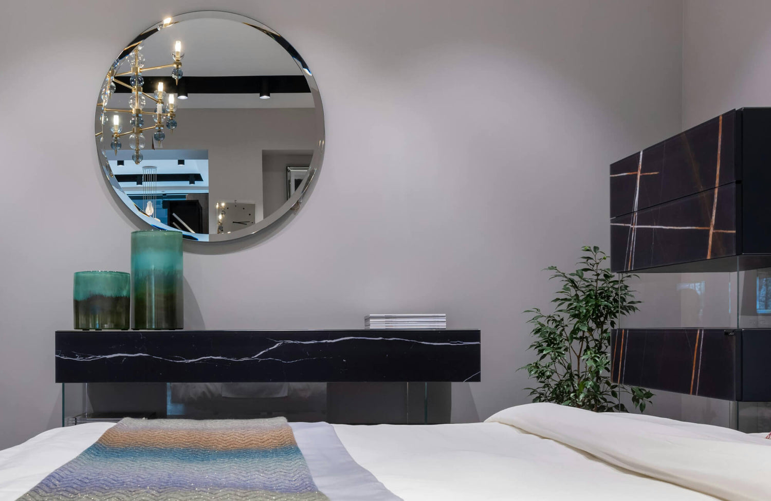 Large round wall mirror centered above a console, reflecting pendant lighting and adding depth to the bedroom space.