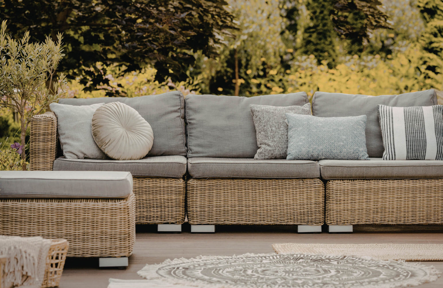 Outdoor wicker sectional sofa with mixed decorative pillows in neutral tones on a sunlit patio surrounded by greenery.