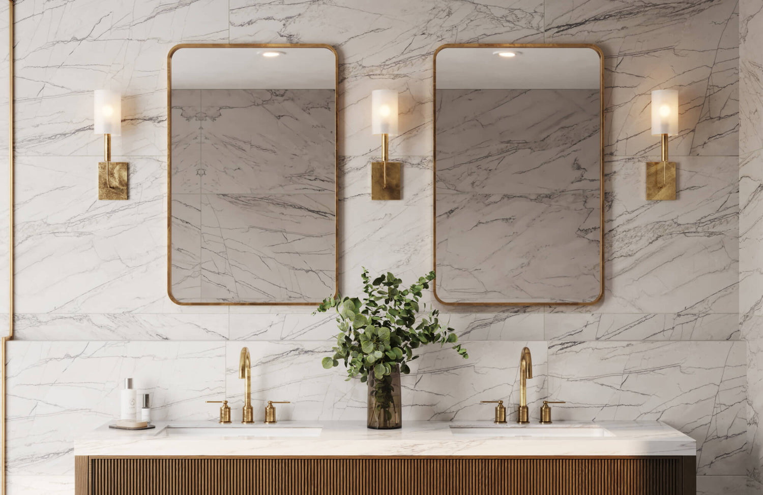 Bathroom featuring dual mirrors with gold frames, marble-look wall tile, brass sconces, and fluted vanity with double gold faucets