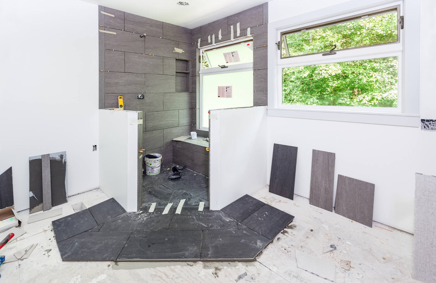A bathroom under renovation with dark grey tiles being installed on the floor and walls, featuring a walk-in shower and a large window.