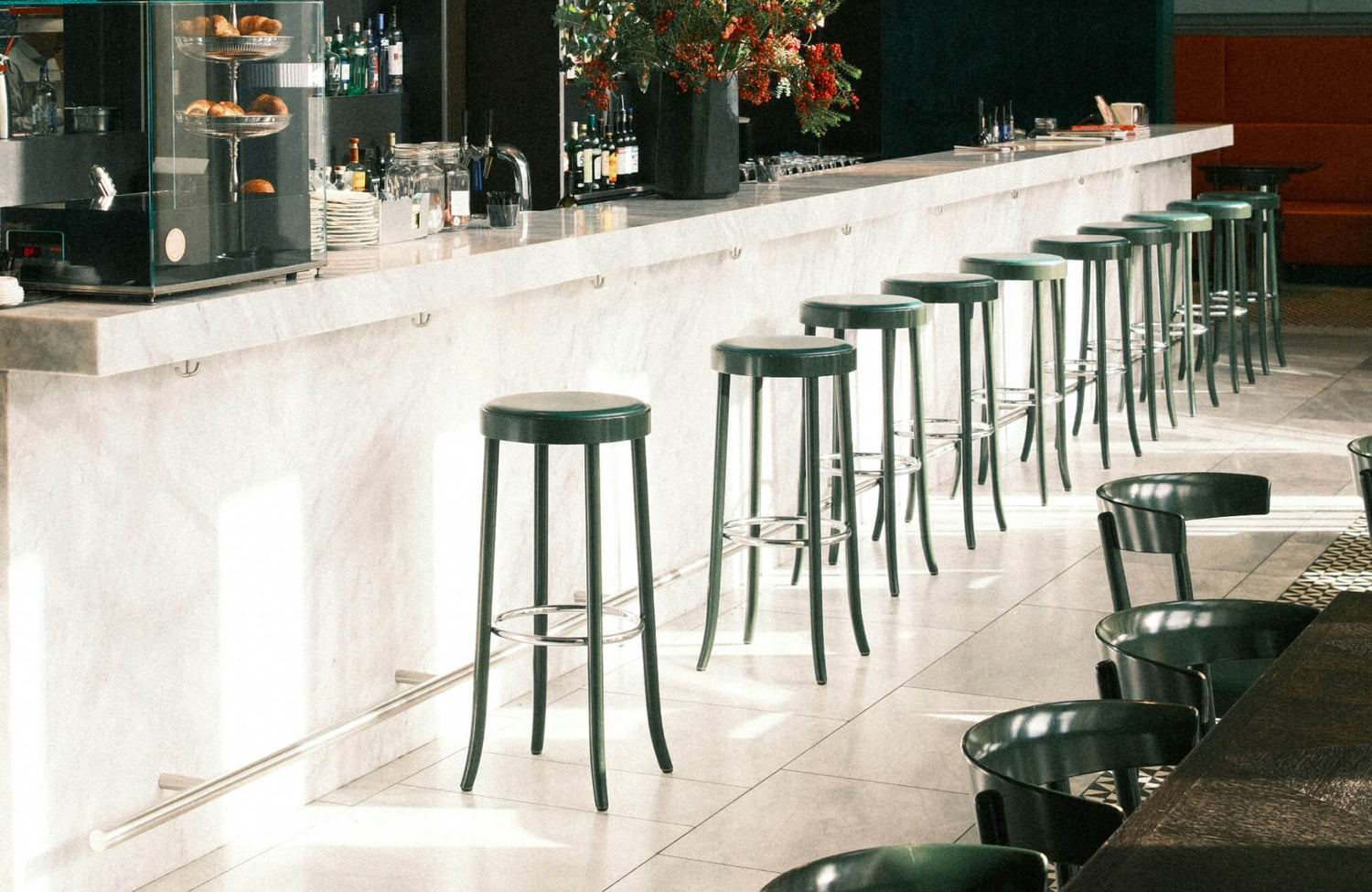 Chic marble bar with sleek black stools, metallic foot rail, and warm morning light casting shadows across the tiled floor.