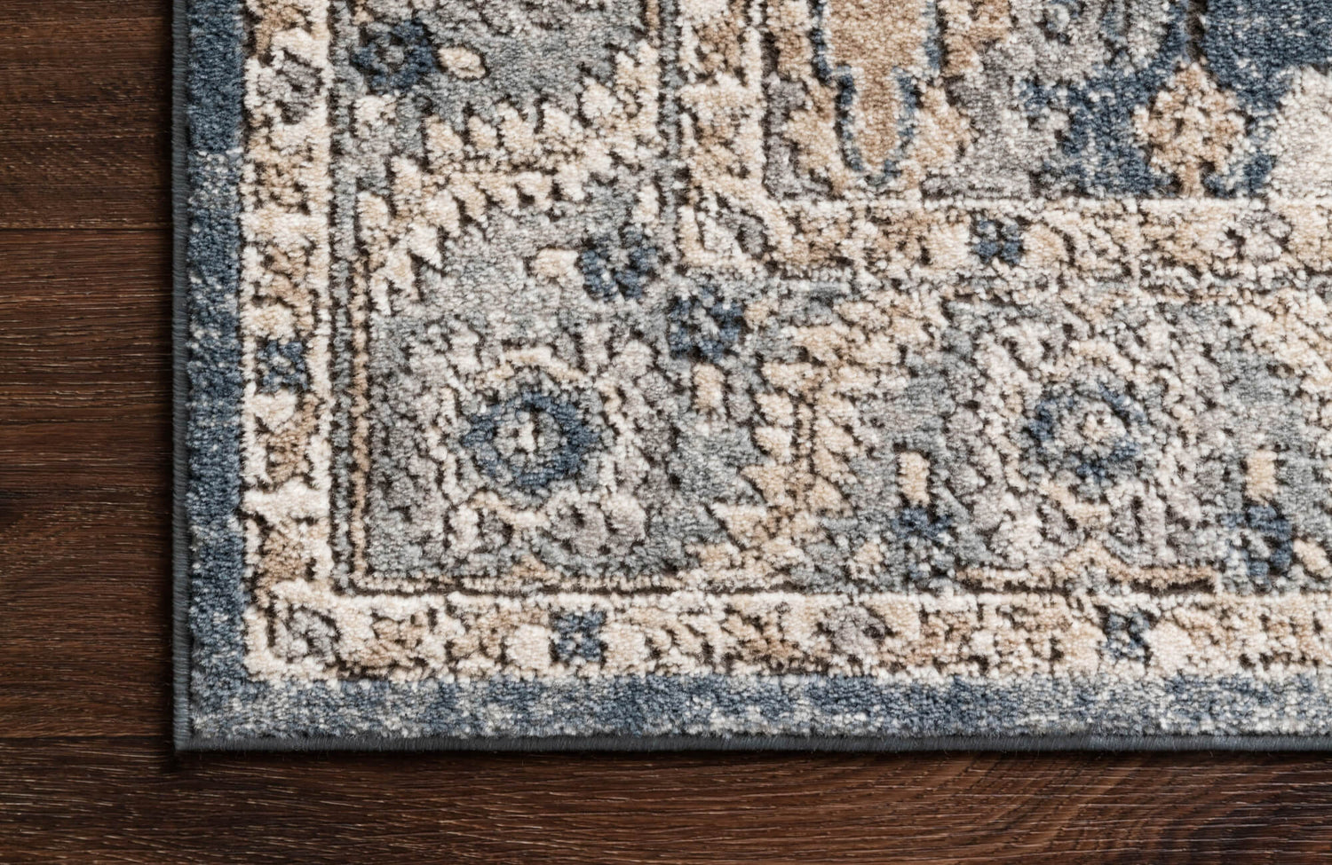 Close-up of a patterned rug in soft gray, beige, and blue tones resting on dark wood flooring, showcasing the detailed border and plush texture.