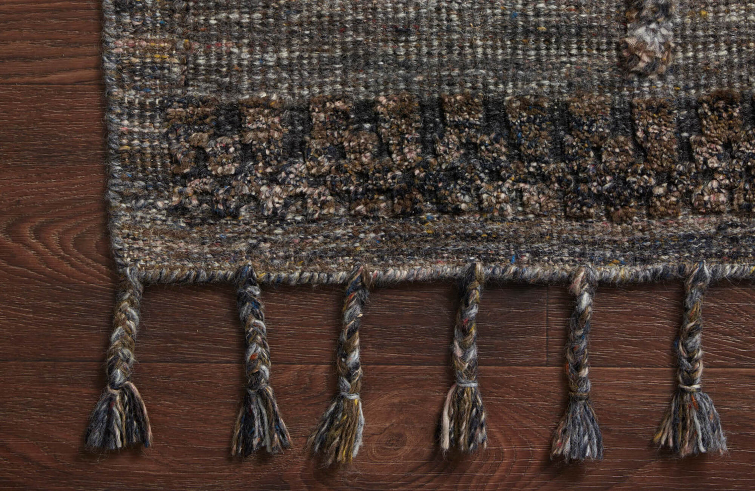 Close-up of a handwoven wool rug with braided fringe details in earthy tones on a rich wood floor.