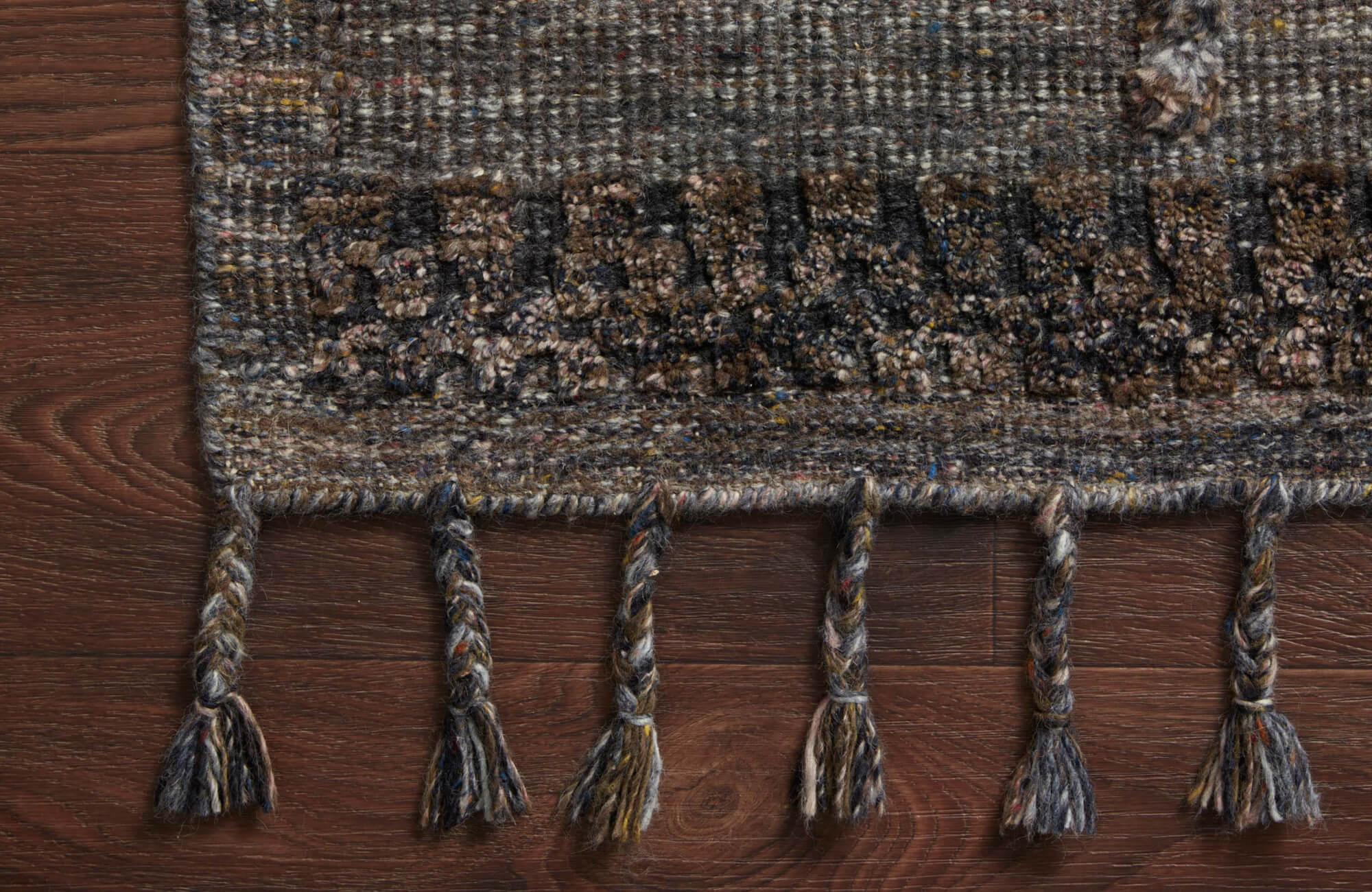 Close-up of a handwoven wool rug with braided fringe details in earthy tones on a rich wood floor.
