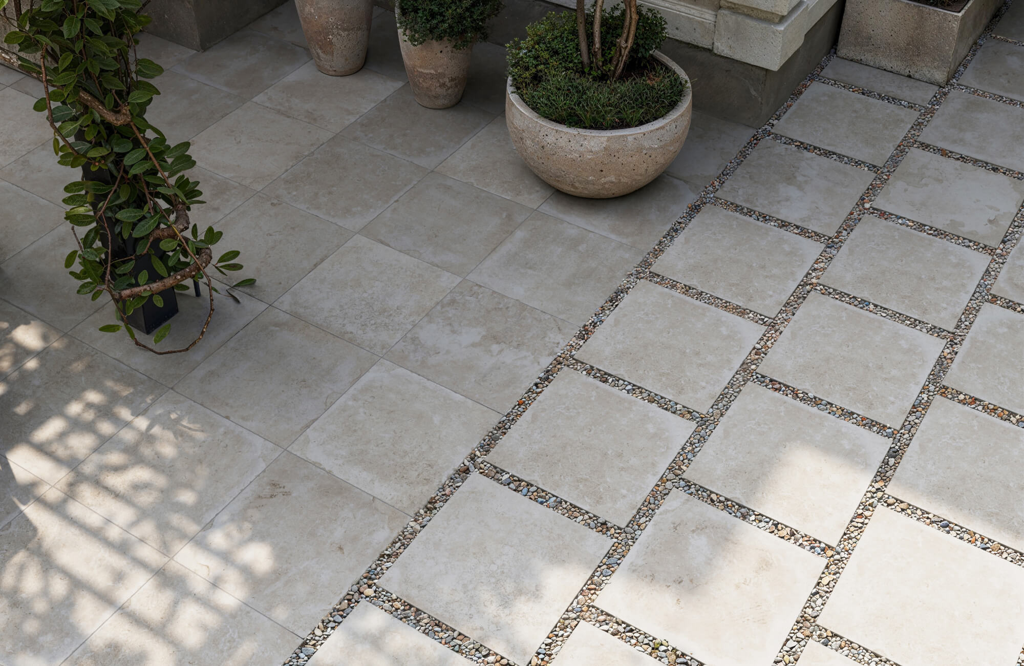Elegant outdoor space with neutral-toned paver tile flooring accented by pebble inlays, naturally illuminated by soft daylight filtering through nearby greenery.