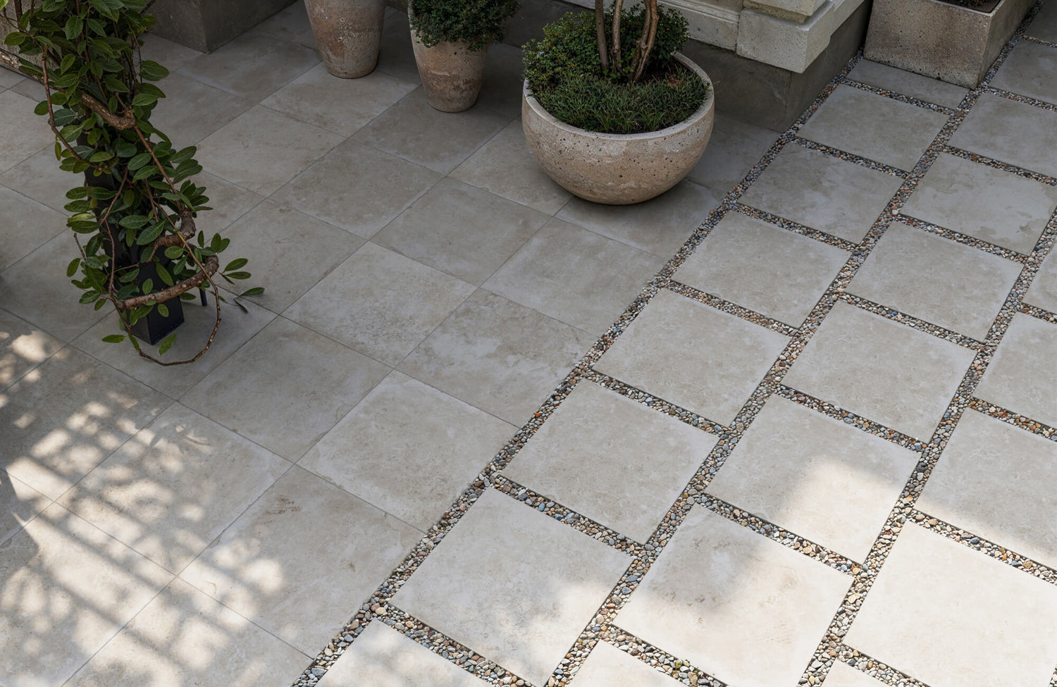 Elegant outdoor space with neutral-toned paver tile flooring accented by pebble inlays, naturally illuminated by soft daylight filtering through nearby greenery.