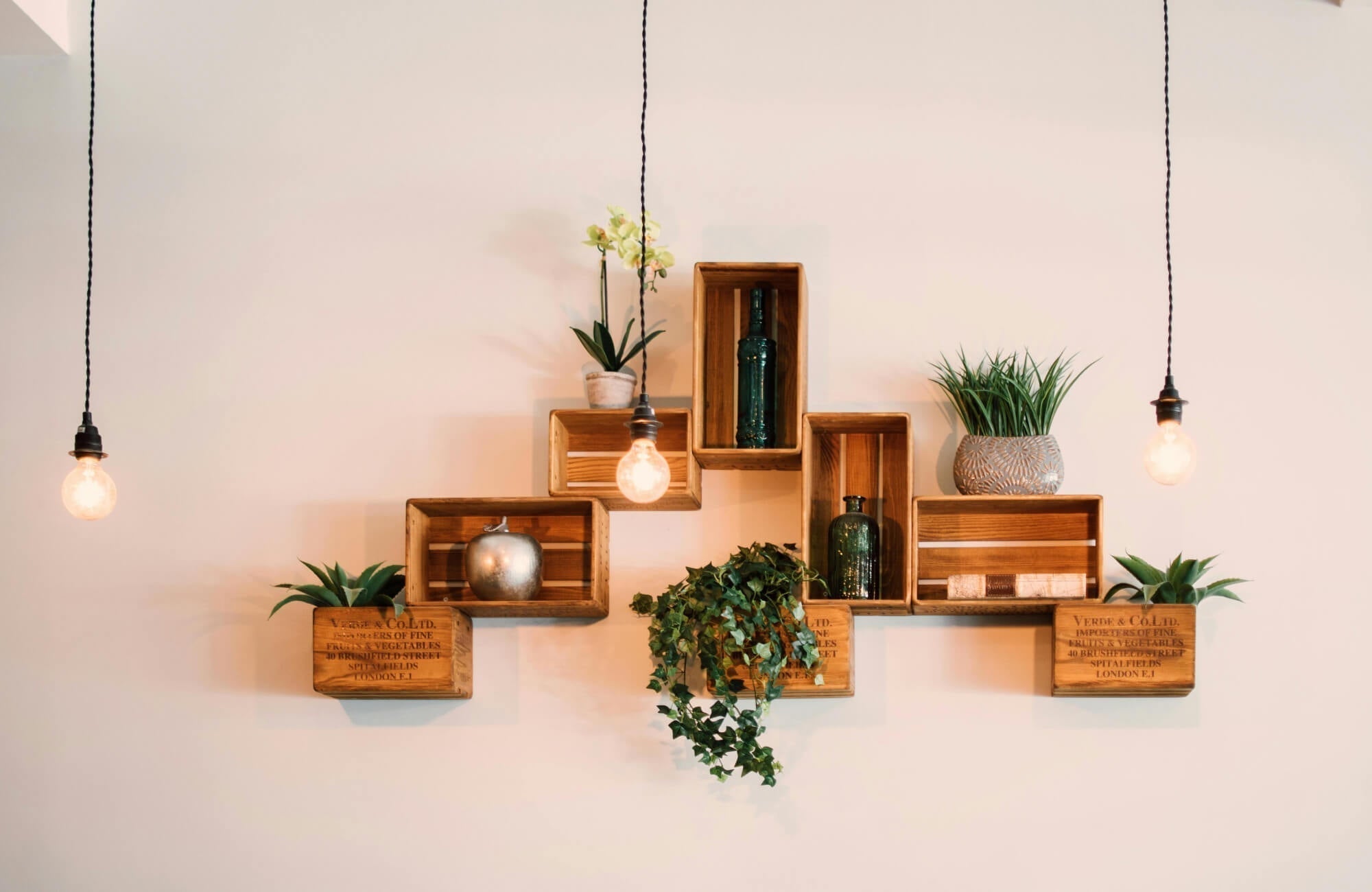Modern wall art made of wooden crate shelves arranged geometrically, styled with plants, glass bottles, and hanging pendant lights.