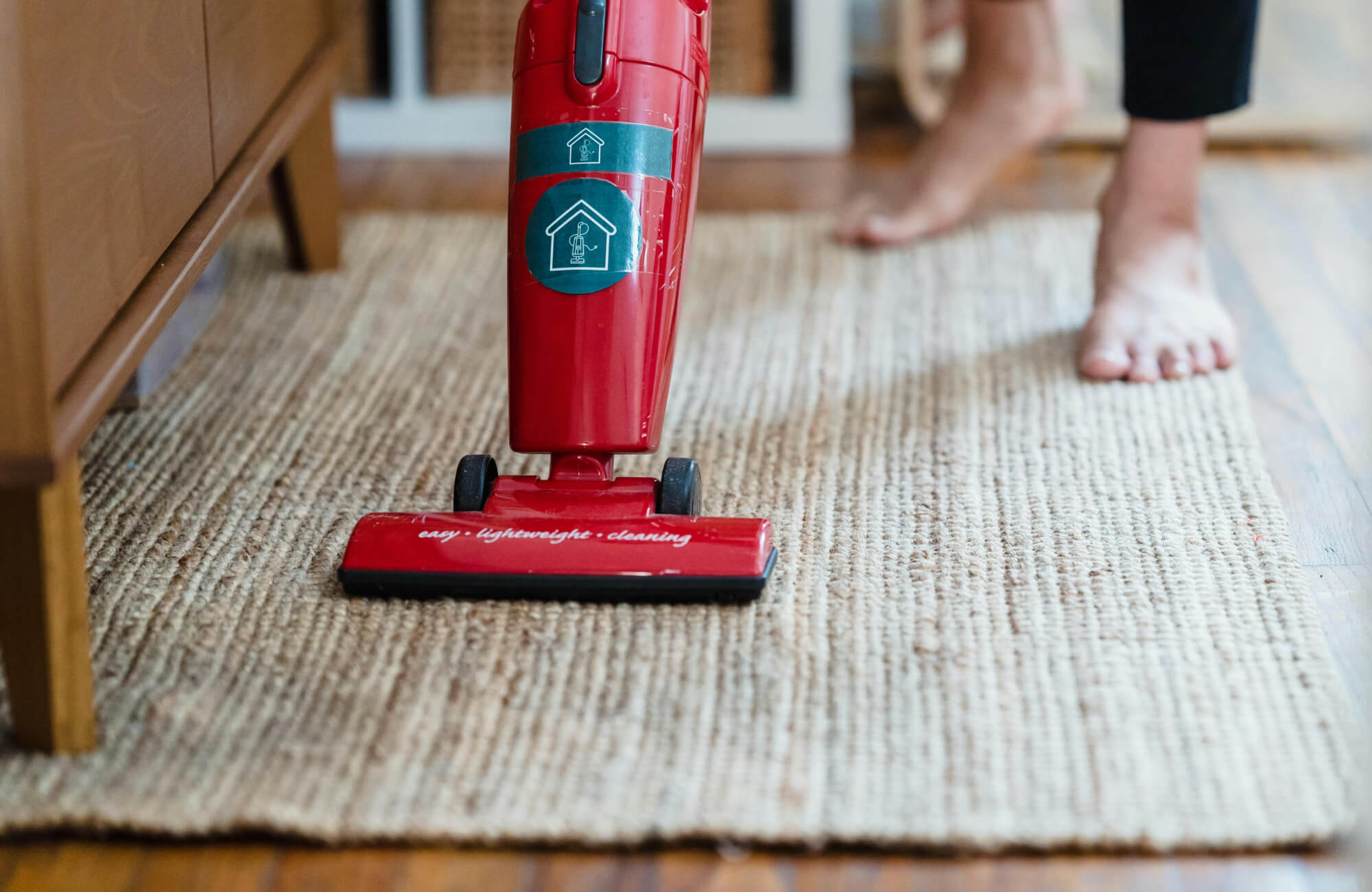 Red upright vacuum cleaning a textured jute rug with bare feet visible in the background.