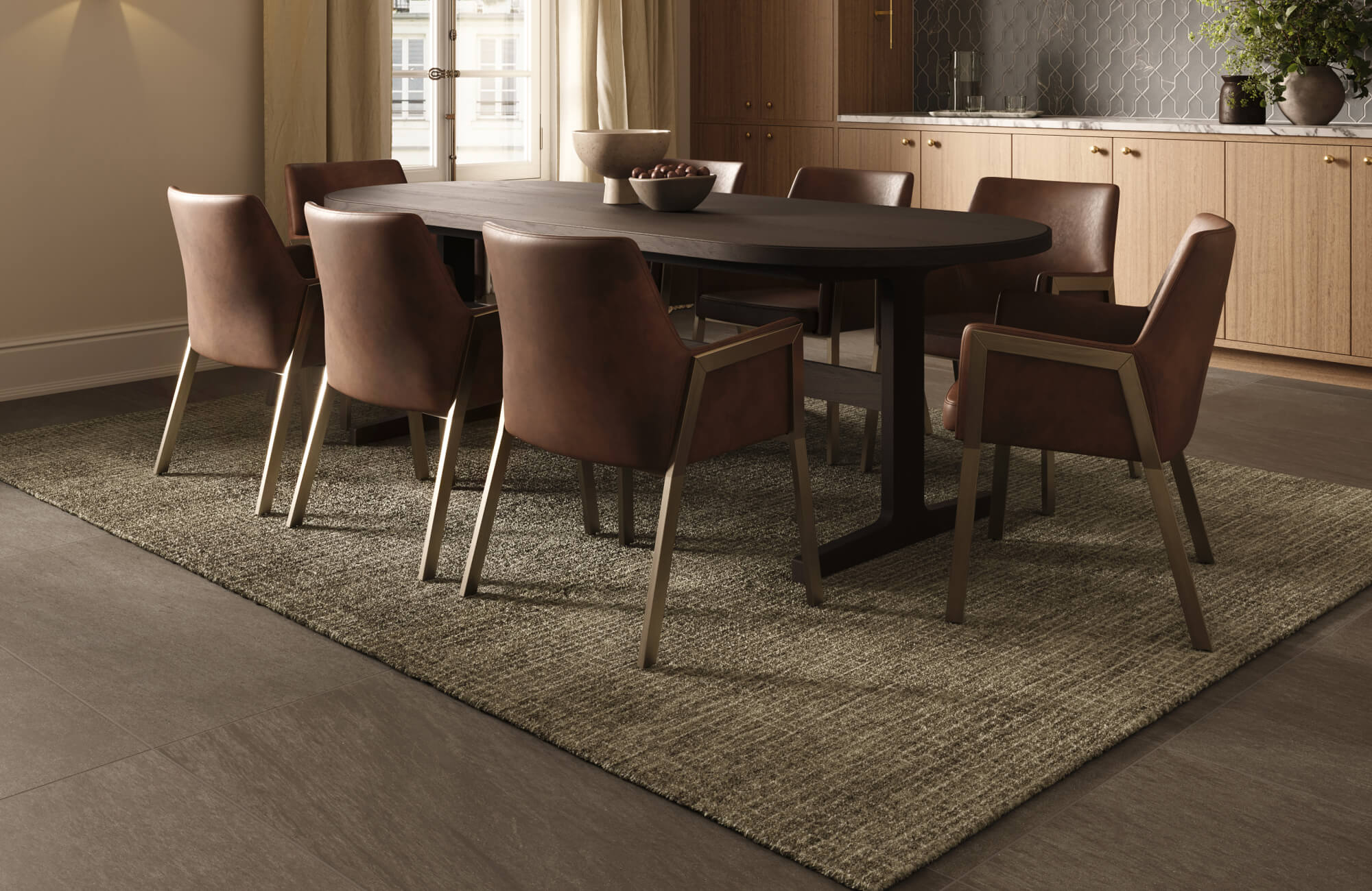 A large brown and beige textured wool rug anchors a dark wood oval dining table with eight brown leather chairs on a dark gray tile floor.