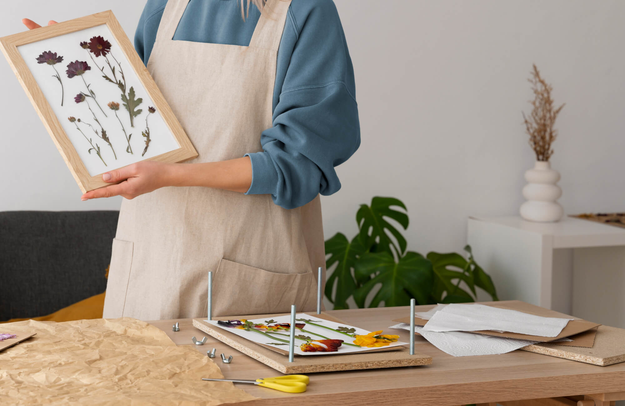 DIY wall art featuring pressed flowers in a wooden frame, held by a person at a crafting table with tools and materials.