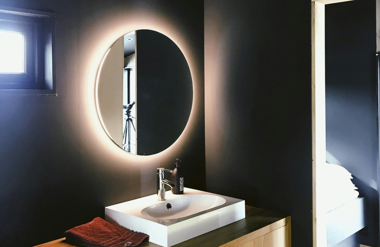 Modern bathroom vanity with a round LED backlit mirror mounted on dark walls, featuring a white basin sink and soft ambient lighting.