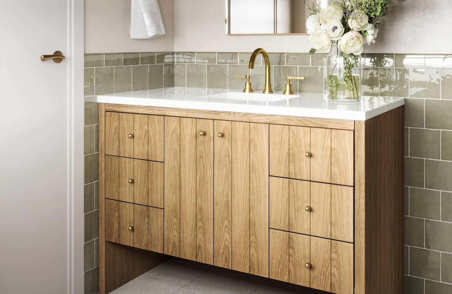 Mid-tone wood bathroom vanity with gold hardware, white sink top, and glass vase of flowers beside a soft green tiled backsplash.