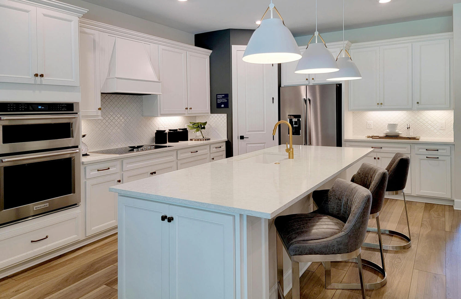 Modern white kitchen featuring layered pendant lighting above a spacious island, paired with brass accents, textured backsplash tile, and soft under-cabinet lighting.