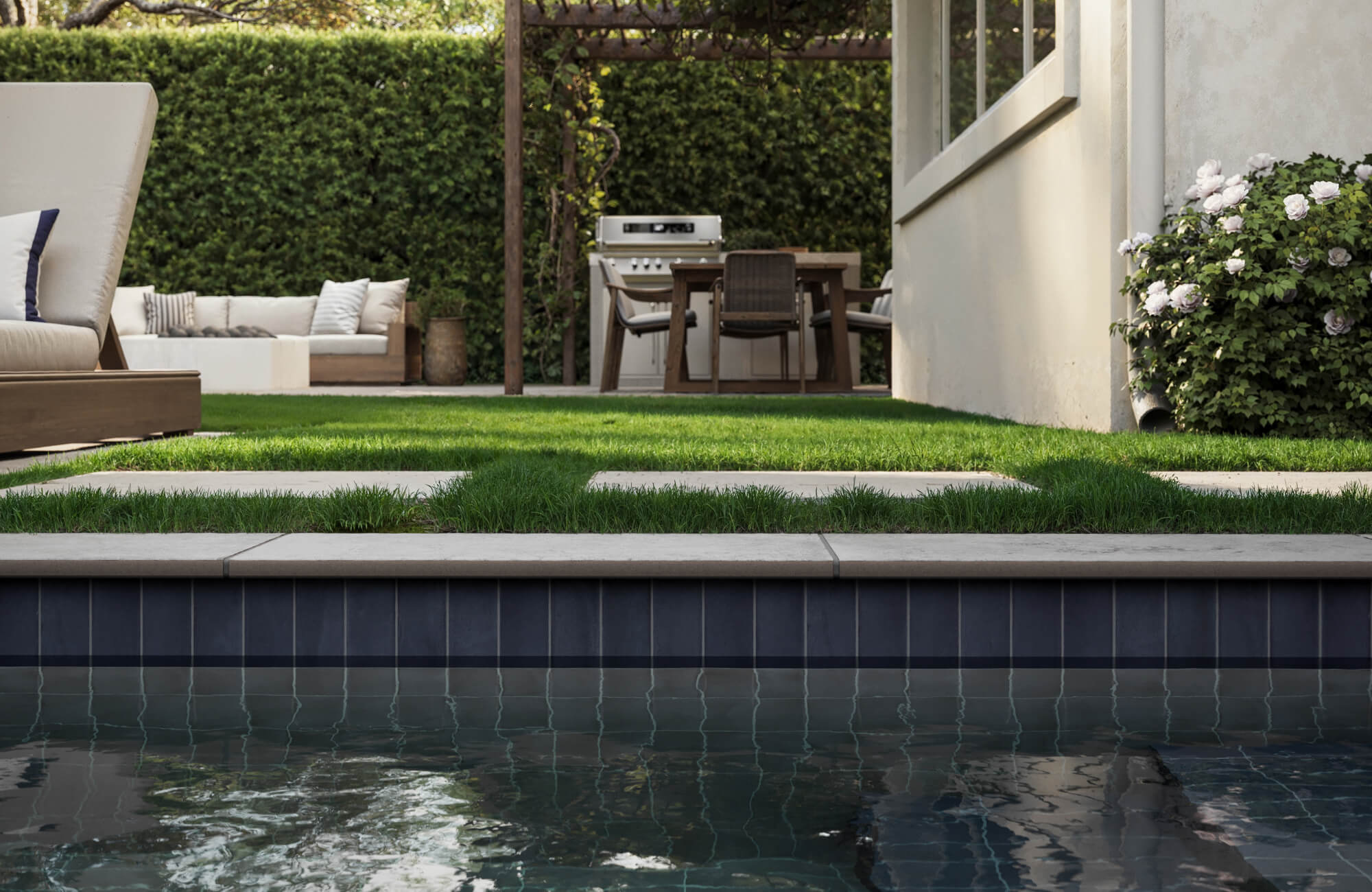 View from a tiled pool toward a landscaped yard with stepping stones, a cozy lounge set, and a wooden pergola dining space.