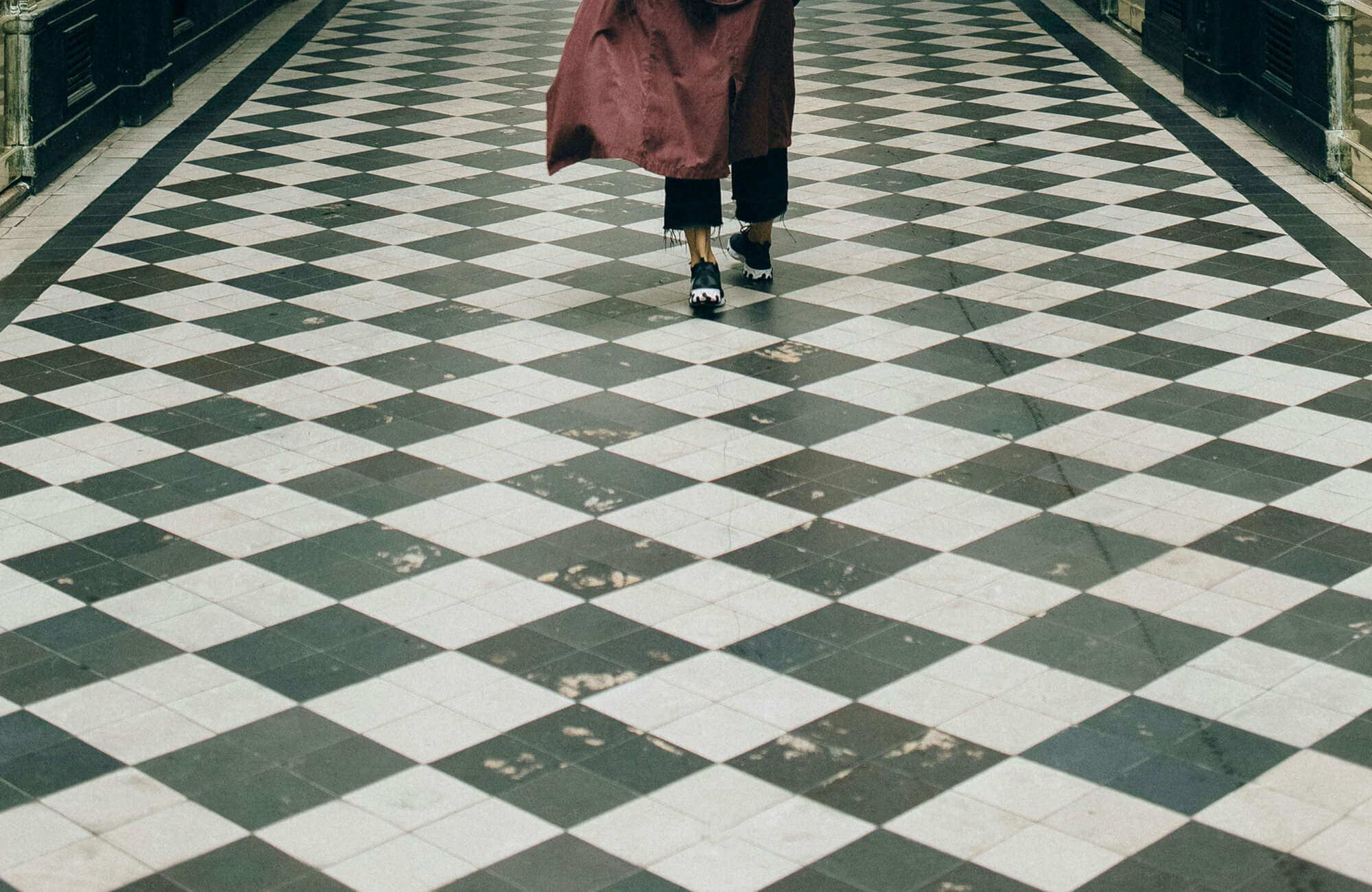 Person walking on a worn black and white checkered tile floor in a covered walkway, with visible scuff marks and surface damage.