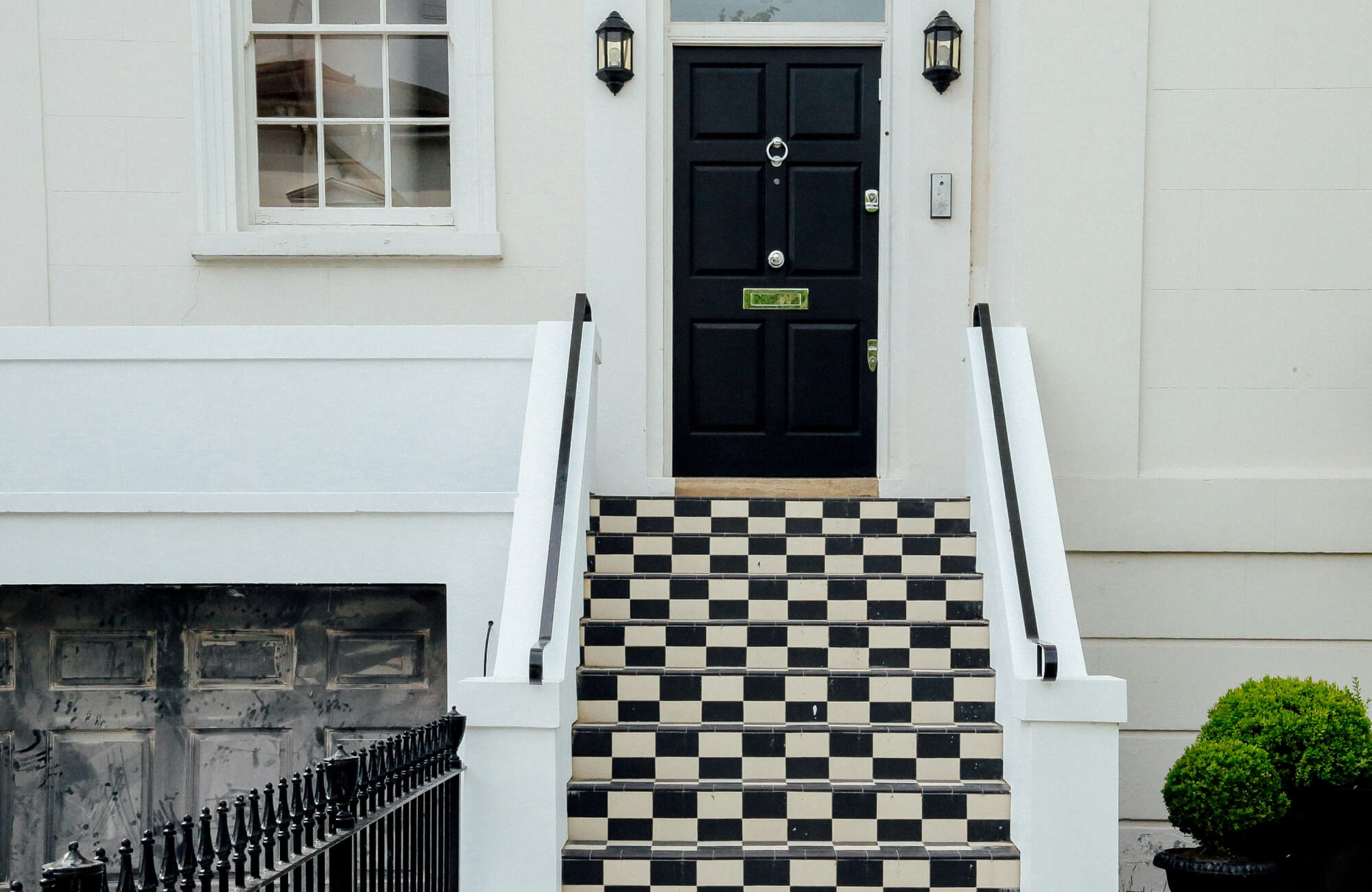 Checkerboard-tiled front steps leading to a black door with white trim on a light-colored townhouse facade.