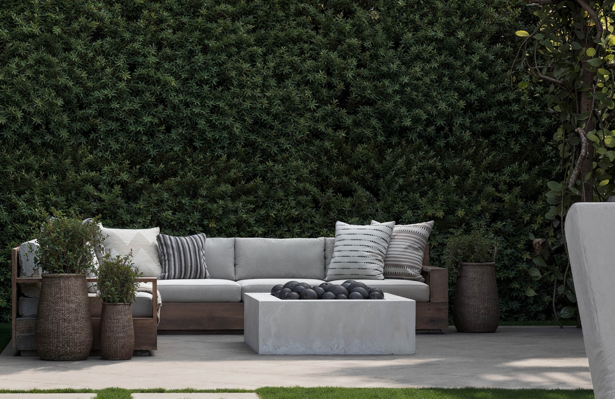 Outdoor garden furniture featuring a modern cushioned sofa with neutral pillows, a concrete coffee table, and decorative planters set against lush greenery in a serene outdoor living space.