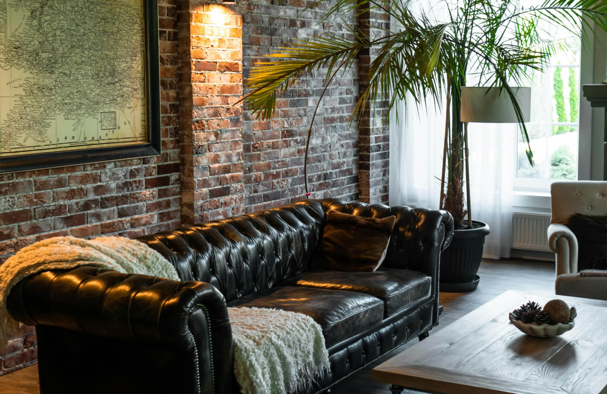 A cozy rustic living room featuring an exposed subway brick wall, a black tufted leather sofa, a wooden coffee table, and a tall potted palm tree.
