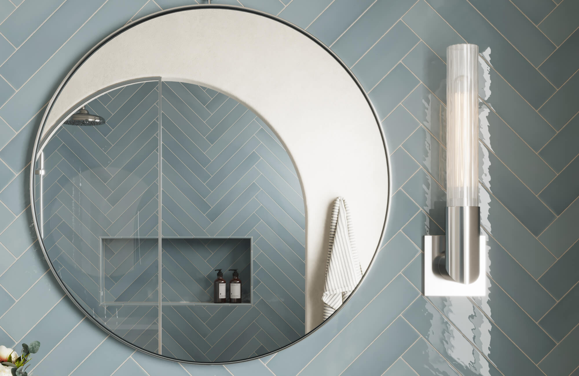 Round silver-framed mirror reflecting a shower with blue herringbone tile walls, built-in niche, and striped towel on a hook.