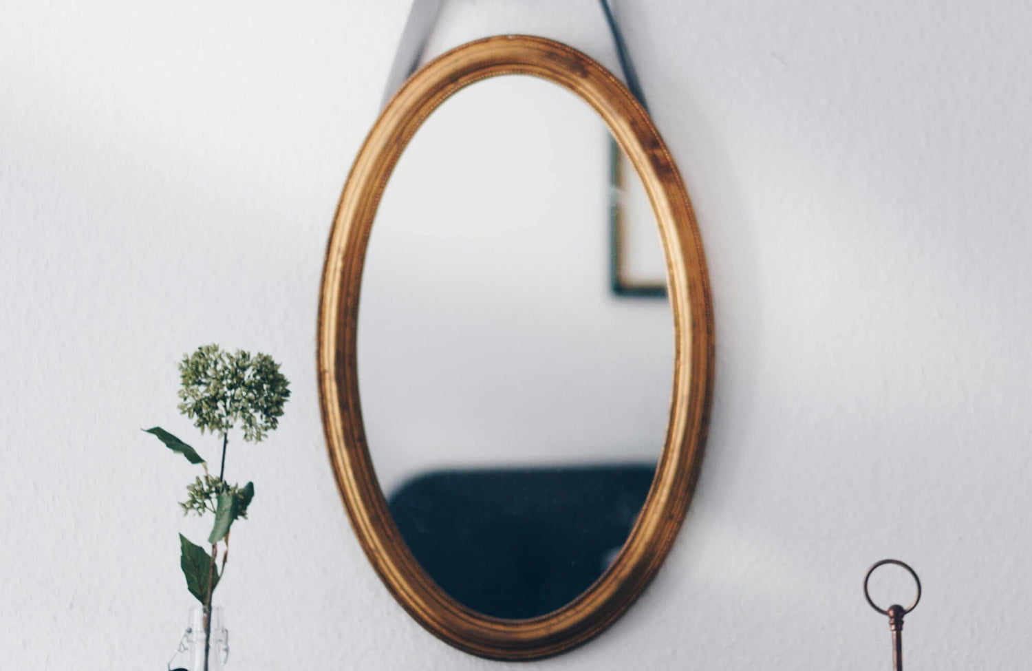 Oval wall mirror with an antique gold frame, suspended by a dark strap above a white wall and a single green flower in a glass vase.
